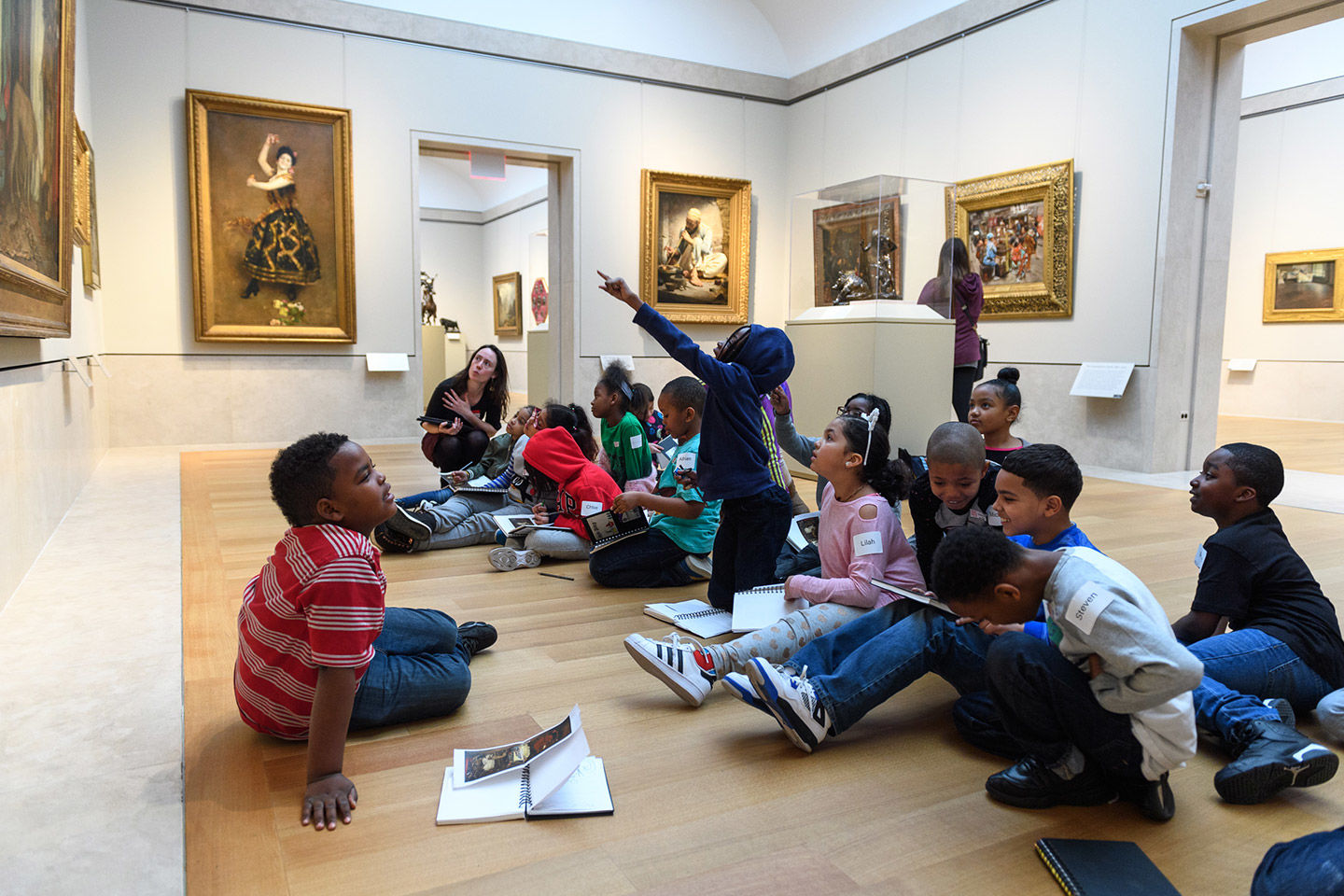 A group of children in the foreground and an adult in the distance sit on a wooden floor of an art gallery; many of the children energetically point up towards something, presumably a work of art, that is not within our view